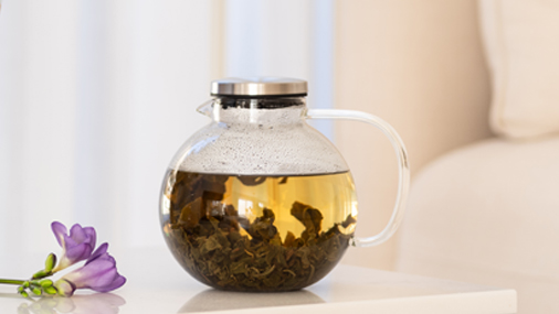 Oolongtee Glaskanne mit Oolong-Teeblättern und Wasser gefüllt auf einem hellen Tisch, daneben liegen lilafarbene Blumen
