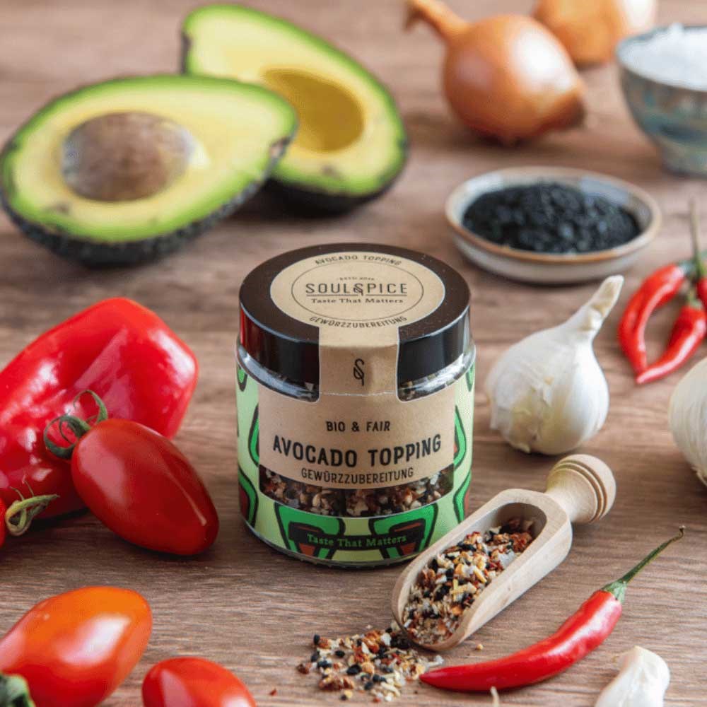 Glas SoulSpice Bio & Fair Avocado Topping Gewürzzubereitung umgeben von Avocado, Tomaten, Paprika, Knoblauch und Chili auf Holztisch