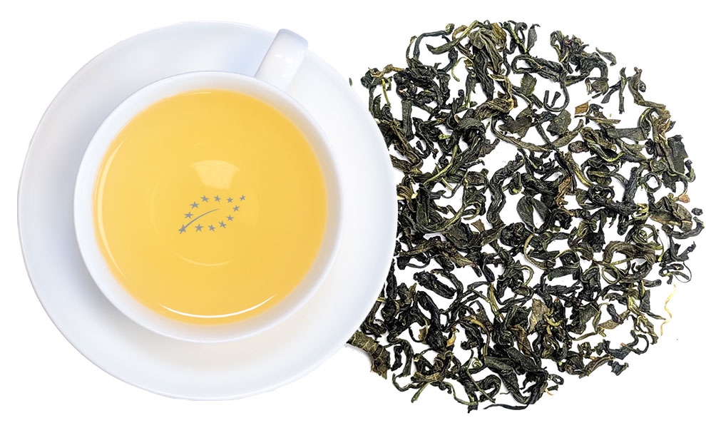 Weiße Tasse mit hellgelbem Grüntee neben losem grünen Teeblatt auf weißem Hintergrund