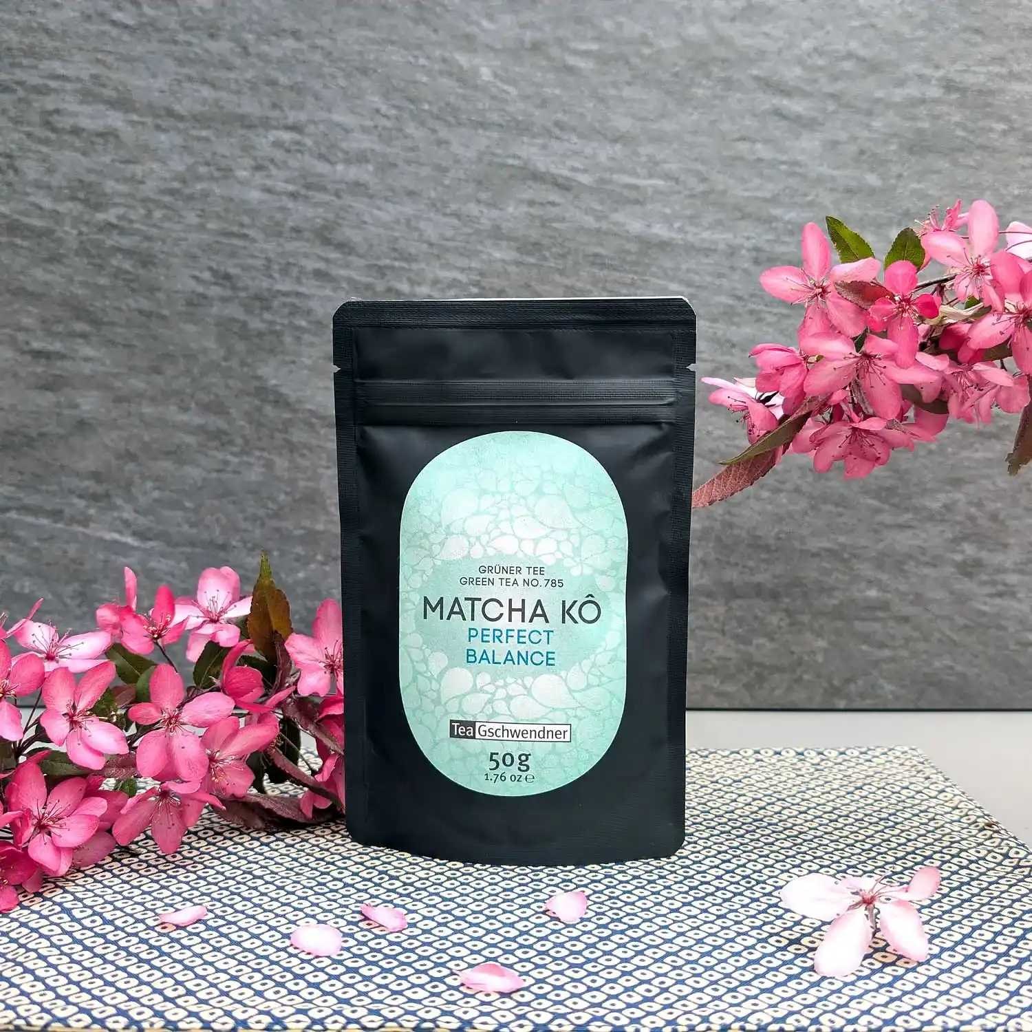 Schwarzer Beutel TeaGschwendner Matcha Kō Perfect Balance, grüner Tee 50 g, dekoriert mit rosa Blüten vor grauem Hintergrund