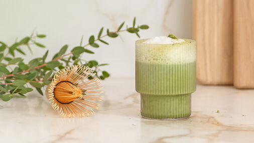 Cremiges grünes Matcha-Getränk im geriffelten Glas mit Milchschaum auf heller Küchenfläche, daneben ein goldener Schneebesen
