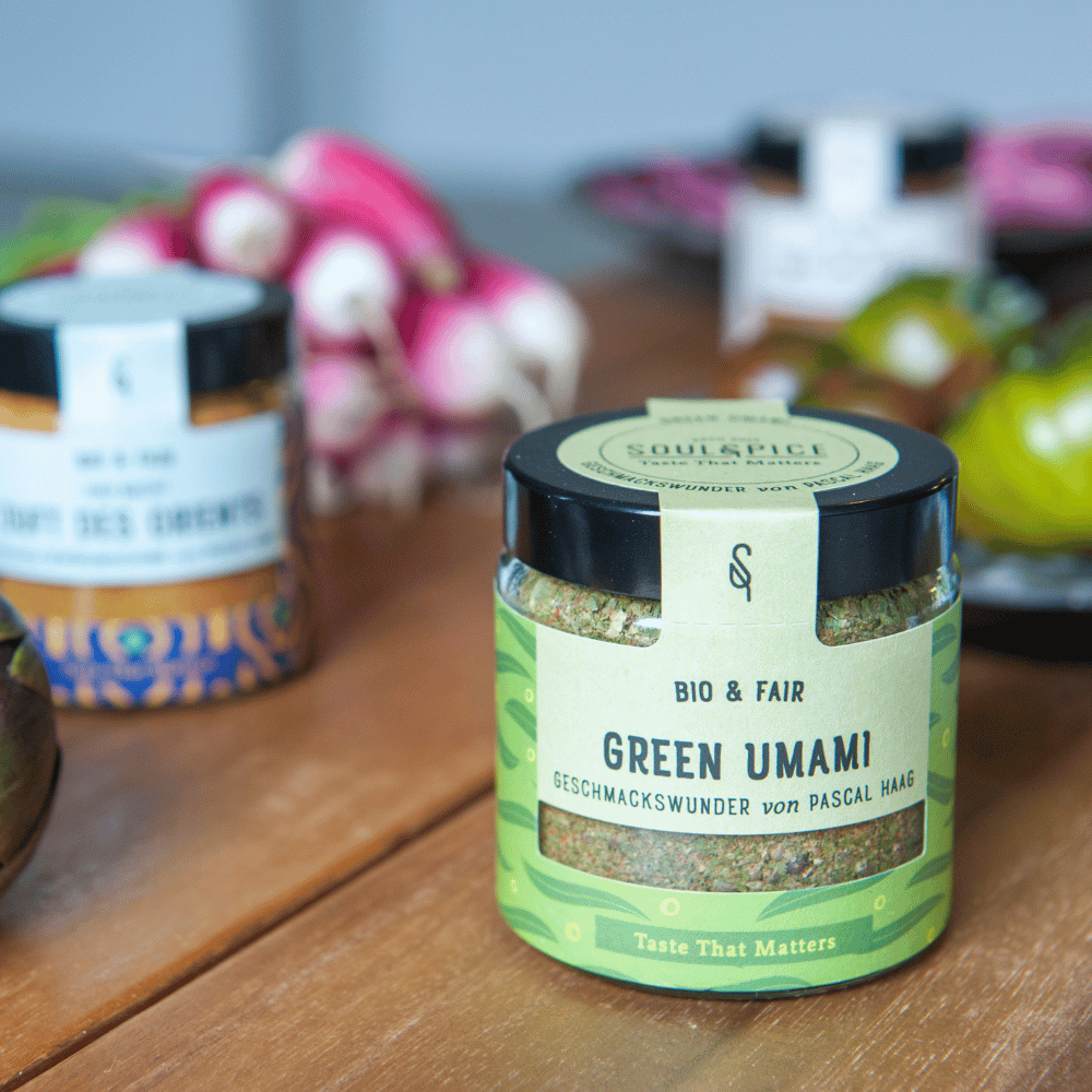 Glas mit SOULSPICE Green Umami Bio & Fair Gewürzmischung von Pascal Haag auf einem Holztisch, im Hintergrund weitere Gewürze und frisches Gemüse