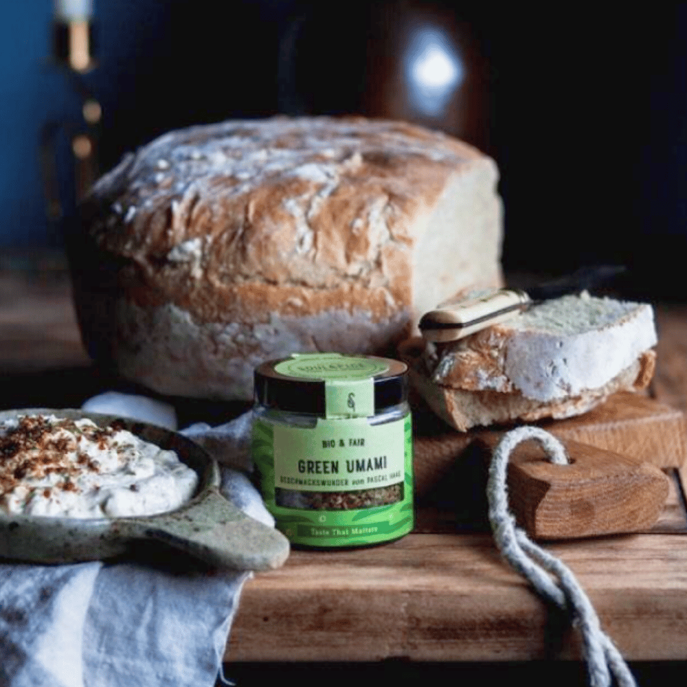 Glas Green Umami Bio-Gewürzmischung vor frischem Bauernbrot mit Brotaufstrich auf einem rustikalen Holztisch