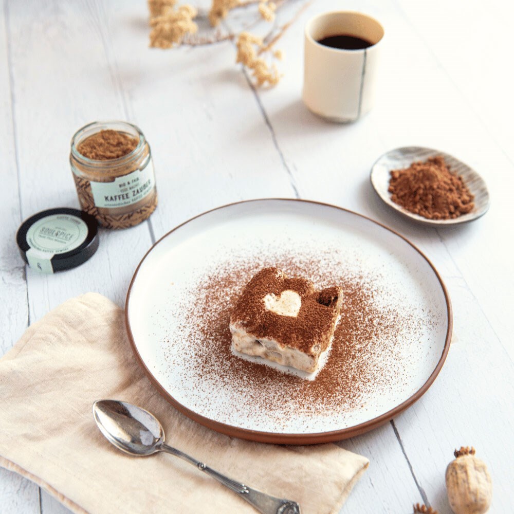 Quadratisches Dessert mit Kakao und Herzdekor auf weißem Teller, daneben Löffel, Tasse Kaffee und Glas Kaffeegewürz auf Holztisch