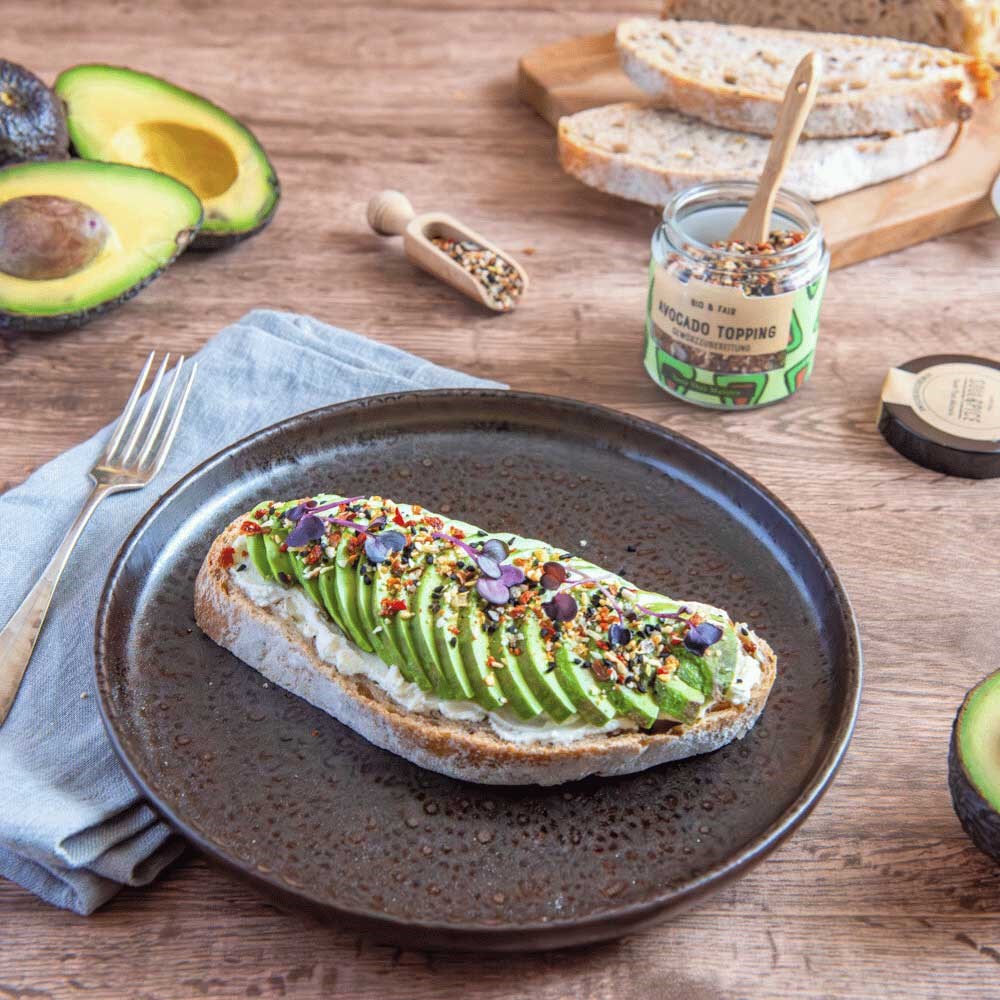 Scheibe rustikales Sauerteigbrot mit Avocadoscheiben, Bio-Avocado-Topping und Kresse auf Keramikteller, daneben frische Avocadohälften und Gewürzglas