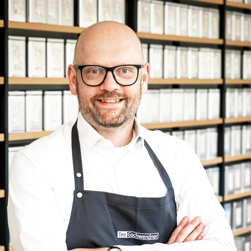 Lächelnder Mann mit Brille und Schürze vor Regalen voller Te